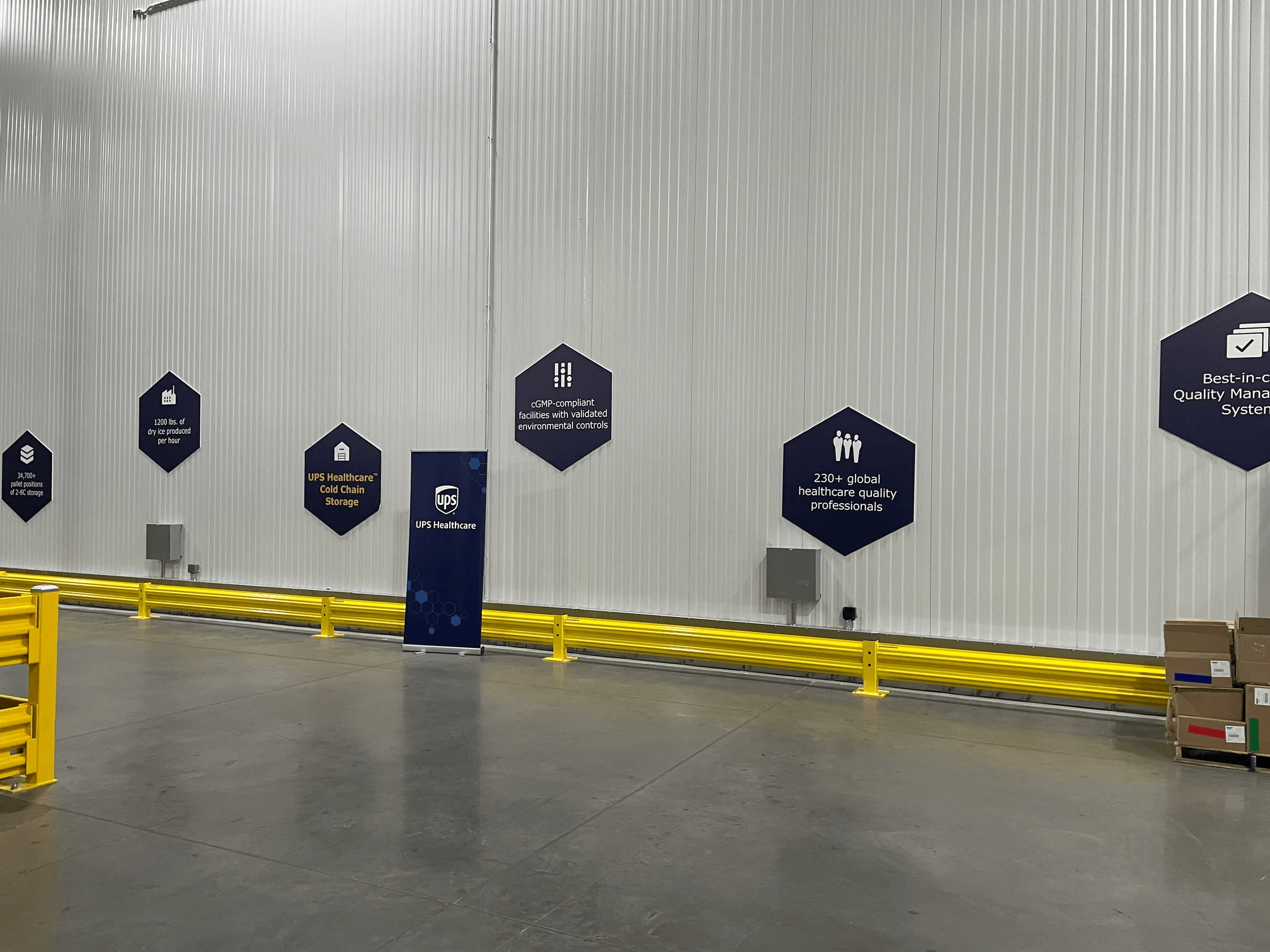 Industrial warehouse interior with UPS Healthcare signs and a banner promoting Cold Chain Storage, safety guardrails in the foreground, and stacks of cardboard boxes to the right.