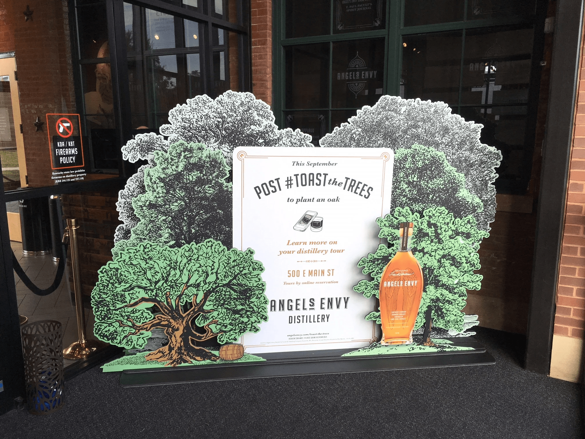 A promotional Angel's Envy whiskey display in front of a brick building features illustrations of green trees and a bottle-shaped cutout with an actual bottle of whiskey. Text on the display promotes an oak planting event.